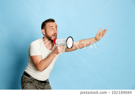 Portrait of emotional bearded man in casual white t-shirt shouting in megaphone against blue studio background Portrait of emotional bearded man in casual white t-shirt shouting in megaphone against blue studio background 102189084