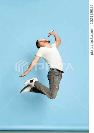 Full-length portrait of bearded energetic man in casual white t-shirt singing in microphone, jumping against blue studio background 102189085