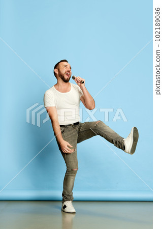 Full-length portrait of bearded happy man in casual white t-shirt singing in microphone, having fun against blue studio background Full-length portrait of bearded happy man in casual white t-shirt singing in microphone, having fun against blue studio background 102189086