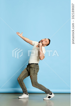 Full-length portrait of bearded funny man in casual white t-shirt singing in microphone, having fun against blue studio background Full-length portrait of bearded funny man in casual white t-shirt singing in microphone, having fun against blue studio background 102189087