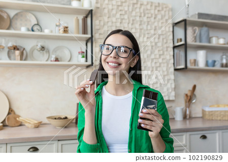 Advertising photo. A young brunette woman in glasses and a green shirt eats a bar of chocolate at home in the kitchen. He looks smilingly and happily at the camera, holding a piece in his hand. Advertising photo. A young brunette woman in glasses and a green shirt eats a bar of chocolate at home in the kitchen. He looks smilingly and happily at the camera, holding a piece in his hand. 102190829