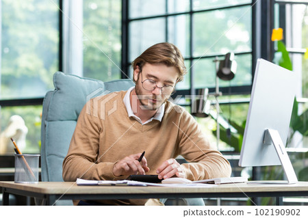 Accounting, audit. A young man works with documents and bills in the office. Calculates and checks reporting, balance, and budget on a calculator. Accounting, audit. A young man works with documents and bills in the office. Calculates and checks reporting, balance, and budget on a calculator. 102190902