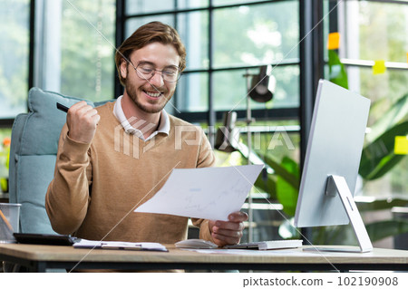 Work in the office. A young man sits at a table with a computer and works with documents, agreements, schedules. He smiles, satisfied with the result. Work in the office. A young man sits at a table with a computer and works with documents, agreements, schedules. He smiles, satisfied with the result. 102190908