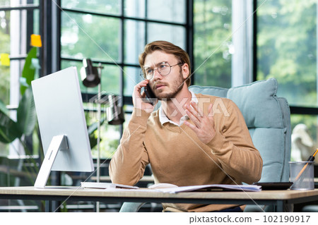 A young man businessman sits in the office at the table and solves questions, problems on the phone. Talks, gesticulates worriedly with his hands, explains. A young man businessman sits in the office at the table and solves questions, problems on the phone. Talks, gesticulates worriedly with his hands, explains. 102190917