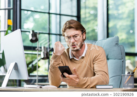 A young man businessman sits in the office at the table and solves questions, problems on the phone. Worriedly looks at the phone, worries, reads messages, checks bills. A young man businessman sits in the office at the table and solves questions, problems on the phone. Worriedly looks at the phone, worries, reads messages, checks bills. 102190926