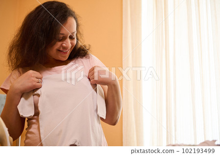 Smiling expectant mother, multi ethnic adult pregnant woman holding ironed newborn clothes, expressing positive emotions during household work in the ironing room. Pregnancy. Motherhood. Maternity Smiling expectant mother, multi ethnic adult pregnant woman holding ironed newborn clothes, expressing positive emotions during household work in the ironing room. Pregnancy. Motherhood. Maternity 102193499