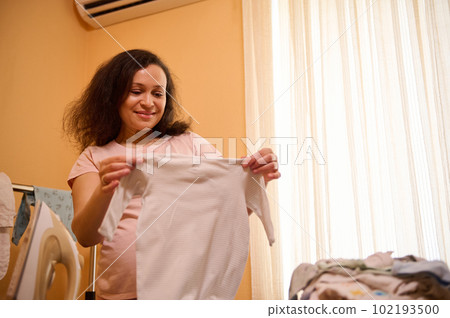 Multi-ethnic delightful happy pregnant adult woman smiling while holding ironed newborn's bodysuit, expressing positive emotions of maternity and pregnancy. People. Lifestyle. Expectation of baby 102193500