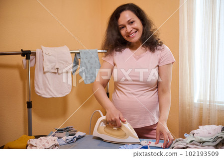 Middle aged multi ethnic positive pregnant woman, wearing pink t-shirt, lovely future mom smiling looking at camera while ironing newborn baby clothes in the home, preparing bag for child birth 102193509