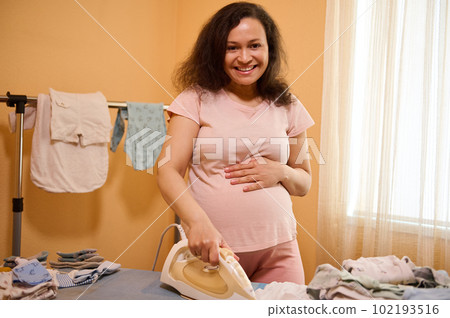 Delightful pregnant adult woman holding hand on her belly, smiles looking at camera while irons clean crumpled newborn clothes. Happy gravid mom, expecting baby, preparing bag for maternity hospital Delightful pregnant adult woman holding hand on her belly, smiles looking at camera while irons clean crumpled newborn clothes. Happy gravid mom, expecting baby, preparing bag for maternity hospital 102193516