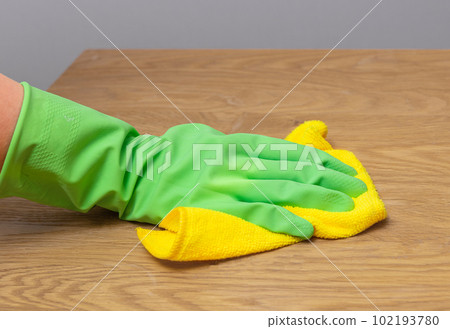 Hand in green glove wiping dust from wooden desk, cleaning wood table with micro fiber cloth, yellow wiper 102193780