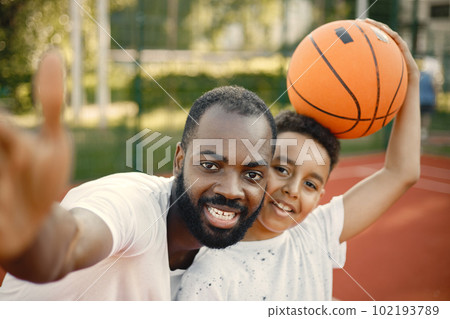 Young father and his son standing on basketball court near the park. Man and boy wearing white t-shirts. Father and son make a selfie. 102193789