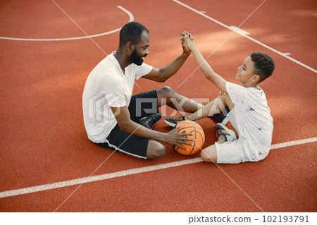 Young father and his son sitting on basketball court near the park. Man and boy wearing white t-shirts. They giving a high five to each other. 102193791
