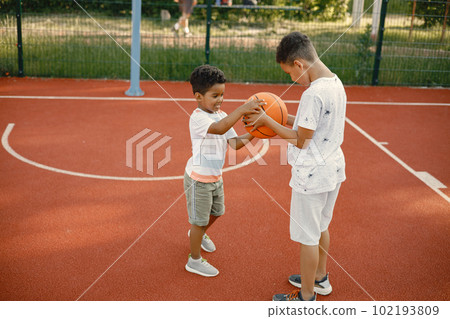 Two multiracional brothers playing basketball on a court near the park. Boys wearing white t-shirts. Older brother teach little one how to play basketball. 102193809