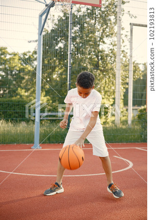 Young boy standing on basketball court near the park. Boy wearing white t-shirt. Boy playing with basketball ball. 102193813