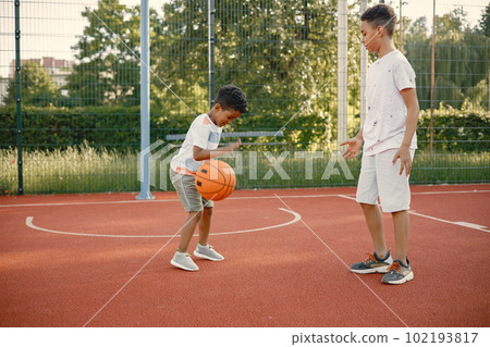 Two multiracional brothers playing basketball on a court near the park. Boys wearing white t-shirts. Older brother teach little one how to play basketball. 102193817