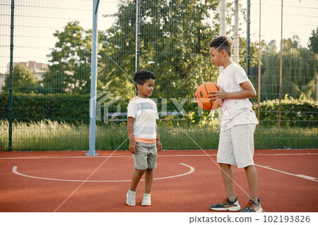 Two multiracional brothers playing basketball on a court near the park. Boys wearing white t-shirts. Older brother teach little one how to play basketball. 102193826