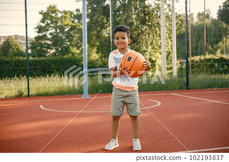Young boy standing on basketball court near the park. Boy wearing white t-shirt. Boy playing with basketball ball. Young boy standing on basketball court near the park. Boy wearing white t-shirt. Boy playing with basketball ball. 102193837