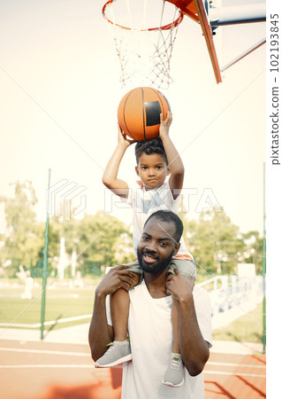 Black father holding on shoulders his little son to help him to score a basket. They standing on a basketball court. Man and boy wearing white t-shirts. Black father holding on shoulders his little son to help him to score a basket. They standing on a basketball court. Man and boy wearing white t-shirts. 102193845