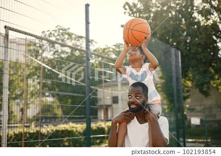 Black father holding on shoulders his little son to help him to score a basket. They standing on a basketball court. Man and boy wearing white t-shirts. Black father holding on shoulders his little son to help him to score a basket. They standing on a basketball court. Man and boy wearing white t-shirts. 102193854