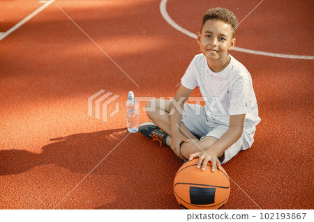 Young boy sitting on basketball court near the park. Boy wearing white t-shirt. Boy has a bottle of water and looking at camera. 102193867