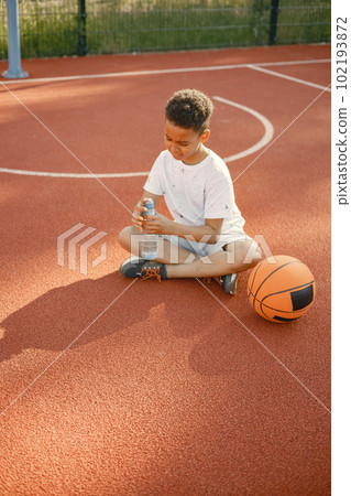 Young boy sitting on basketball court near the park. Boy wearing white t-shirt. Boy holding a bottle of water. 102193872