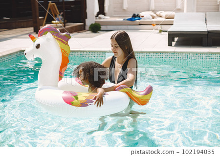 Curly multiracial girl and her caucasian mother playing in the pool. Girl wearing yellow swimwear and mother wearing a black one. Woman and girl have big unicorn swim ring. 102194035