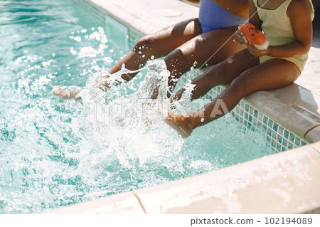 Two multiracial sisters sitting near the pool. Girls wearing blue and yellow swimwear. Girls splashing the water in pool and holding a peace of watermelon. 102194089