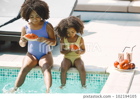 Two curly multiracial sisters sitting near the pool. Girls wearing blue and yellow swimwear. Girls eating a peace of watermelon. 102194105