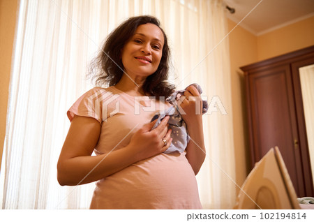 Gravid woman, expectant mother is waiting and preparing for the birth of child at home, holding ironed baby bodysuit, expressing positive emotions of maternity and pregnancy, smiling looking at camera Gravid woman, expectant mother is waiting and preparing for the birth of child at home, holding ironed baby bodysuit, expressing positive emotions of maternity and pregnancy, smiling looking at camera 102194814
