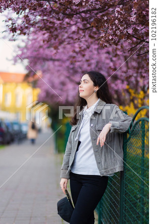Young smiling Caucasian woman wearing jacket posing at street. Blooming tree in background. Vertical orientation. Concept of carefree and psychology Young smiling Caucasian woman wearing jacket posing at street. Blooming tree in background. Vertical orientation. Concept of carefree and psychology 102196182