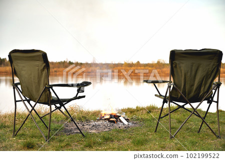 A pair of tourist armchairs, a folding chair, a bonfire on the river bank in the evening at sunset, twilight in autumn at sunset, relaxation and rest, the beginning of autumn, tourism and travel, a 102199522
