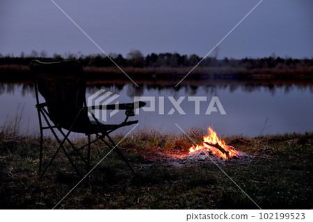 Tourist chair, folding chair, bonfire on the river bank in late evening, twilight in the fall at sunset, relaxation and rest, early autumn, close-up Tourist chair, folding chair, bonfire on the river bank in late evening, twilight in the fall at sunset, relaxation and rest, early autumn, close-up 102199523