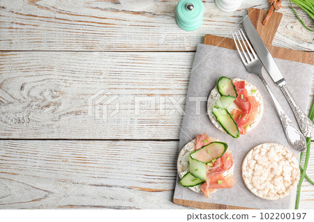 Puffed rice cakes with prosciutto and cucumber served on white wooden table, flat lay. Space for text 102200197