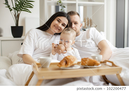 Baby drinking from sippy cup while parents snuggling at home 102200477