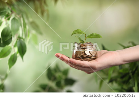 Woman holding coins and green sprout on blurred background, closeup. Money savings 102200512