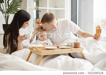 Parents with baby having meal in bed of studio apartment 102200526