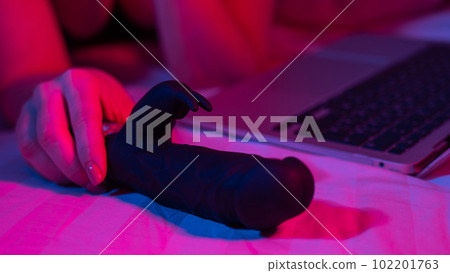 A woman lies in bed with a black dildo and looks at a laptop. Girl using sex toy in blue-red light.  102201763