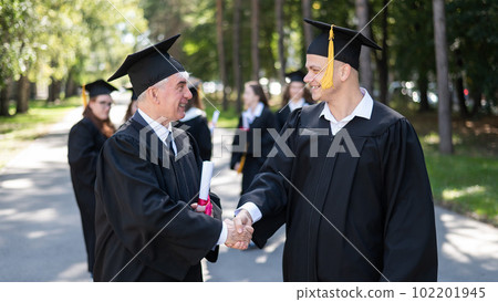 A group of graduates in robes outdoors. An elderly man and a young guy congratulate each other on receiving a diploma. 102201945