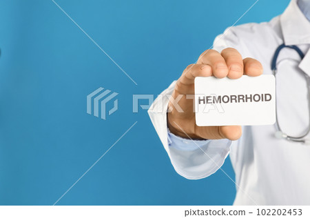 Doctor holding business card with word HEMORRHOID on blue background, closeup. Space for text 102202453