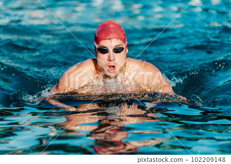 Swimming man - male swimmer swimming breaststroke. Portrait of man doing breast stroke swimming in pool outside wearing red swimming cap and swim goggles. Fit athlete training. Swimming man - male swimmer swimming breaststroke. Portrait of man doing breast stroke swimming in pool outside wearing red swimming cap and swim goggles. Fit athlete training. 102209148