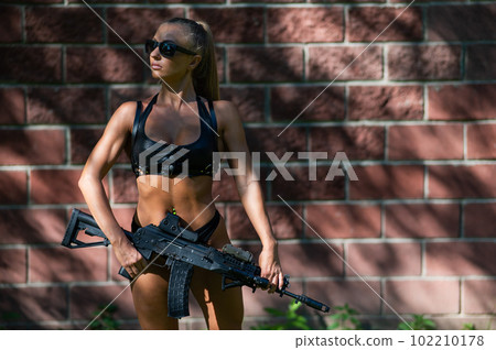 A caucasian woman in a bikini holds a machine gun against a brick wall. 102210178
