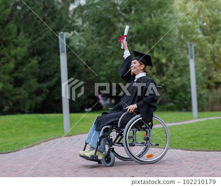Caucasian woman in a wheelchair in a graduate costume rejoices at receiving a diploma. Caucasian woman in a wheelchair in a graduate costume rejoices at receiving a diploma. 102210179