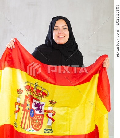 Balanced smiling Muslim woman in traditional black hijab holds flag of Spain Balanced smiling Muslim woman in traditional black hijab holds flag of Spain 102210809
