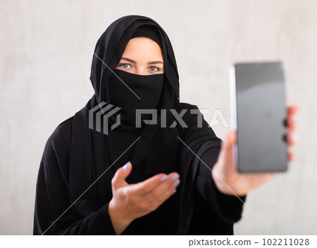 young Muslim girl with her face covered by burka shows empty dark smartphone screen. young Muslim girl with her face covered by burka shows empty dark smartphone screen. 102211028