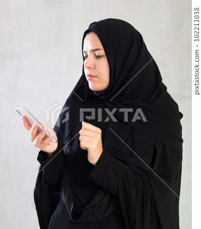 Worried young woman dressed in traditional islamic clothes looking up something on her phone 102211038