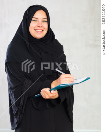 gleeful girl in hijab holds several notebooks and writes important note in her diary gleeful girl in hijab holds several notebooks and writes important note in her diary 102211040