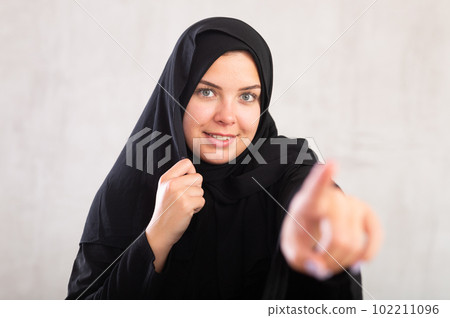 Cheerful young muslim lady in black traditional hijab pointing to side Cheerful young muslim lady in black traditional hijab pointing to side 102211096