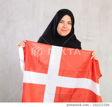Balanced smiling Muslim woman in traditional black hijab holds flag of Denmark Balanced smiling Muslim woman in traditional black hijab holds flag of Denmark 102211104