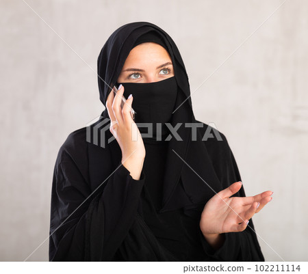 light-skinned Muslim woman with covered face with black niqab is talking on mobile phone light-skinned Muslim woman with covered face with black niqab is talking on mobile phone 102211114