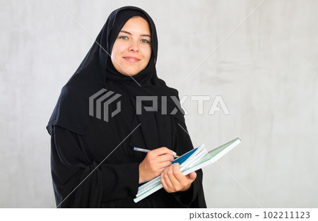 gleeful girl in hijab holds several notebooks and writes important note in her diary 102211123